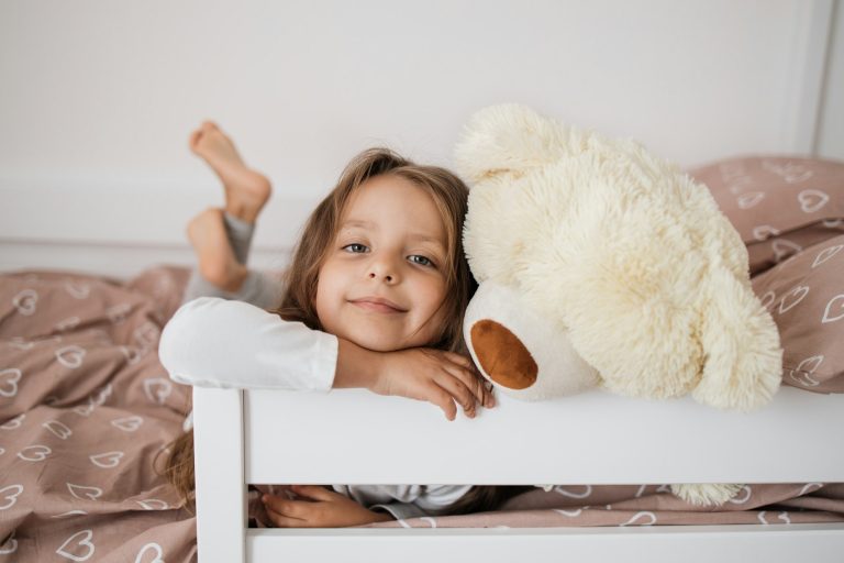 Health and beauty concept - little girl with teddy bear lying on bed at home.